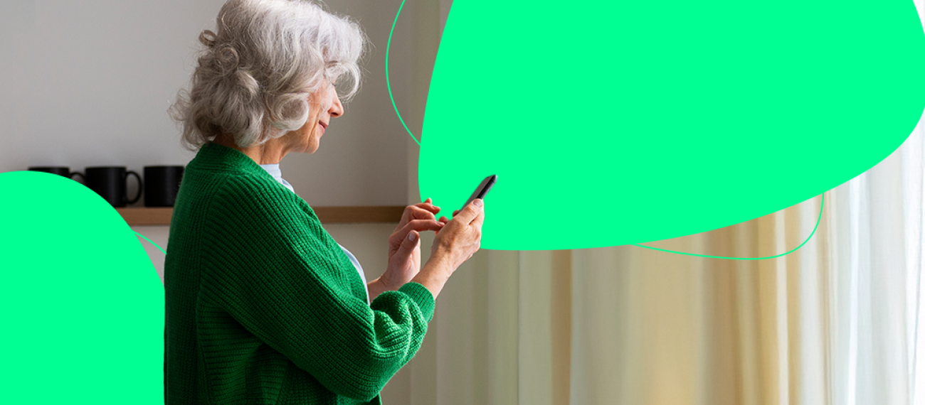 Ältere Frau mit grauen Haaren nutzt ein Smartphone vor einem Fenster mit Vorhängen – Symbolbild für digitale Gesundheitsanwendungen und einfache Medikamentenbestellung per App.